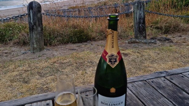 A bottle of Bollinger champagne and two glasses on a wooden table with a view of the beach and ocean during sunset. - Roberts Beach Escape in Tairua is 3-5 minute walk from the beach (Tairua)
