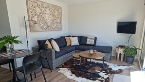 A modern living room with a sectional sofa, a coffee table, a dining area with a chair, a flat-screen TV mounted on the wall, and a large decorative wall piece. - Roberts Beach Escape in Tairua is 3-5 minute walk from the beach (Tairua)