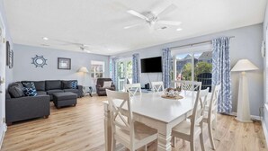 A spacious living room with a grey sectional sofa, a white dining table with chairs, a ceiling fan, and a TV mounted on the wall. - Seabreeze Getaway (5BR Beachfront Home w/ Pool) (Surfside Beach)