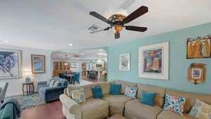 A living room with a large beige sectional sofa, a blue armchair, and a coffee table. There is a ceiling fan with a decorative light fixture, and artwork on the walls.