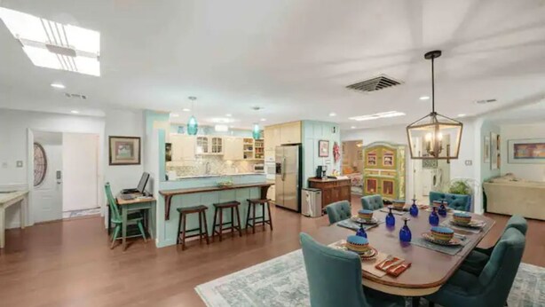 A spacious living area with a large wooden dining table, blue chairs, and a kitchen in the background.