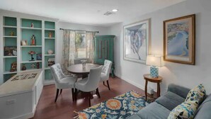 A dining area with a round table and chairs, a teal-colored cabinet with shelves, a window with curtains, and a painting on the wall.