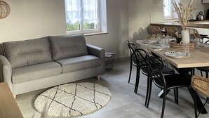 A modern living room with a grey sofa, a dining table set for four, and a large window with sheer curtains.