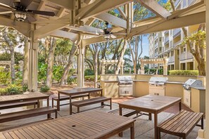 Un patio al aire libre con mesas y bancos de madera, un ventilador de techo y una zona de barbacoa.