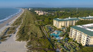 An aerial view of a coastal resort with beachfront access, multiple hotel buildings, and a central pool area surrounded by greenery.