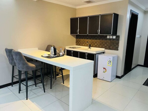 A compact kitchen with a white countertop, black cabinets, a small refrigerator, and two grey upholstered chairs. - Asala AL Shorouk Serviced Apartments (Al Khobar)