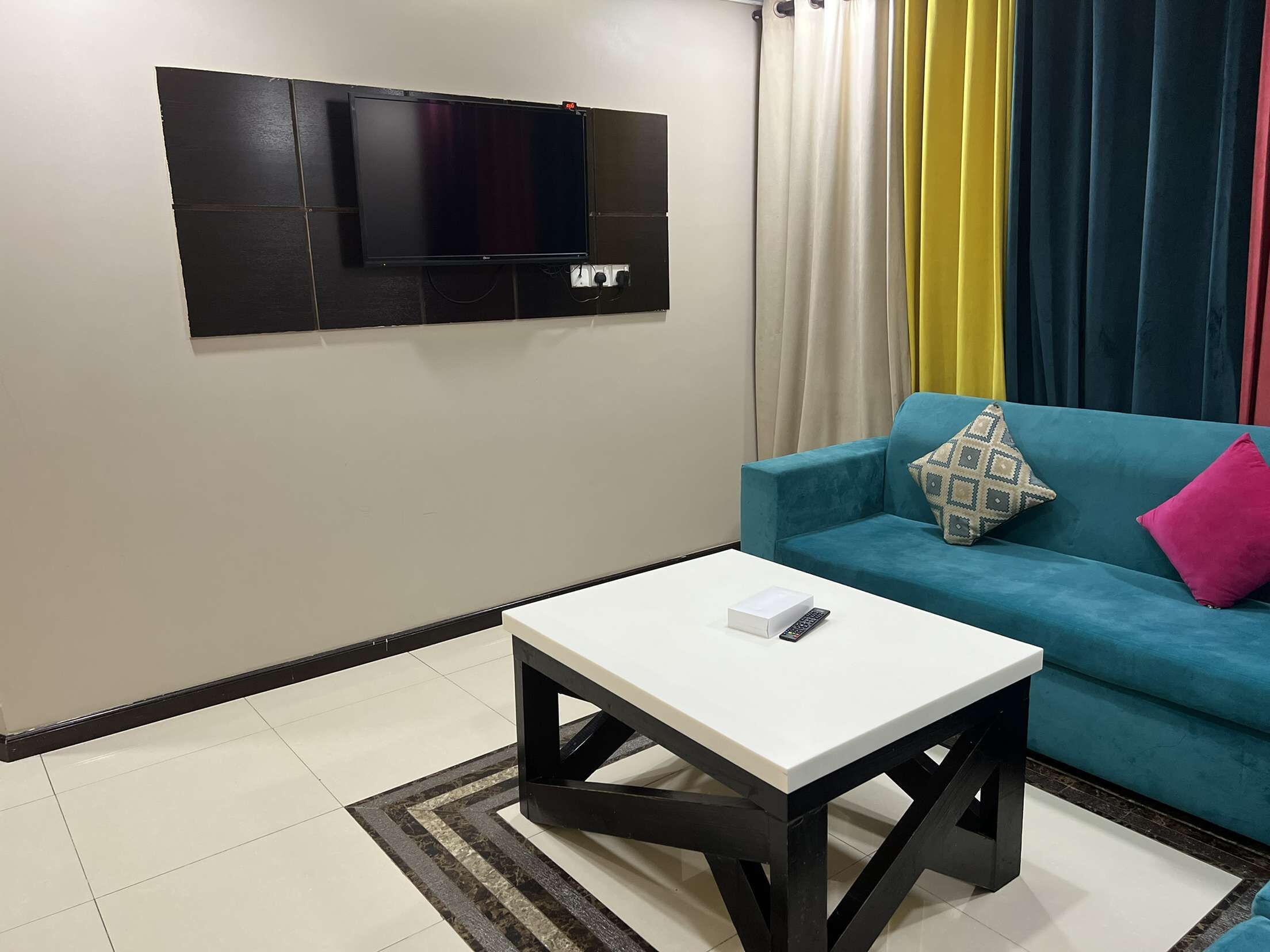 Een moderne woonkamer met een flatscreen televisie aan de muur, een blauwe bank met gekleurde kussens, een witte salontafel en een gestreept vloerkleed.