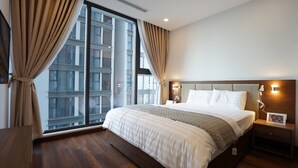 Moderni hotellihuone, jossa on suuri sänky, puinen pääty ja näkymä kaupungin siluettiin lattiasta kattoon ulottuvien ikkunoiden kautta.