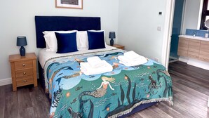 A bed with a blue headboard, two bedside tables with lamps, a framed picture on the wall, and a bathroom visible through an open door. - The Long Barn, Brick Kiln Barns, Dilham - Sleeps 6 in Luxury - Dog Friendly (Dilham)