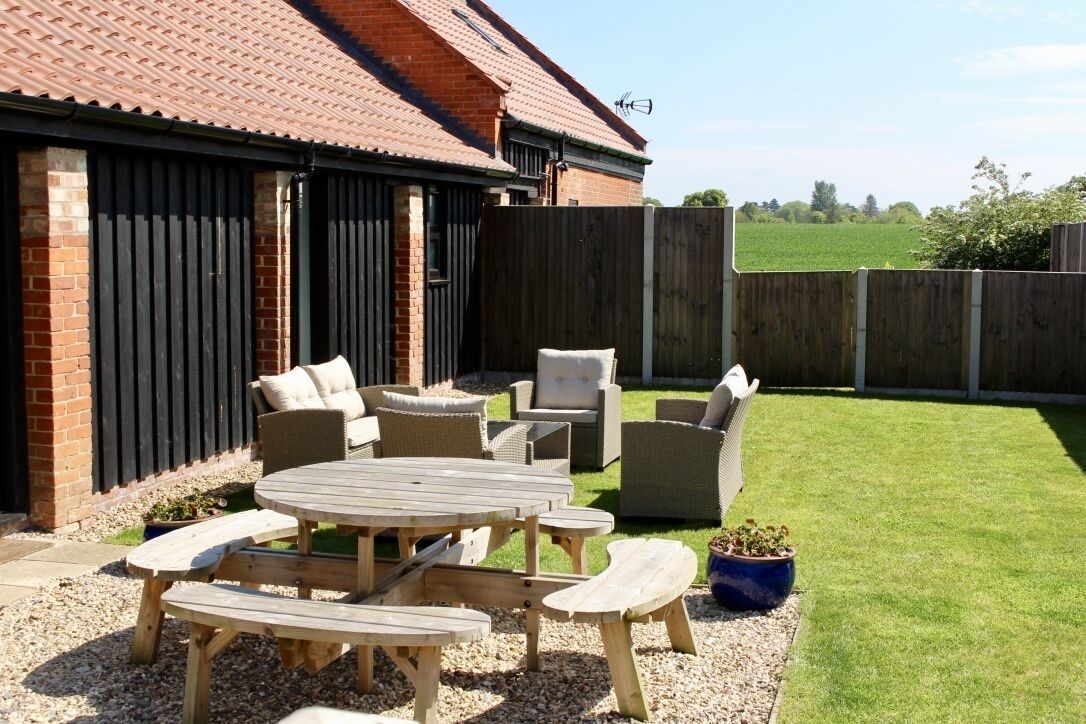 A patio area with a wooden table and chairs, a brick building with a tiled roof, and a fenced garden.