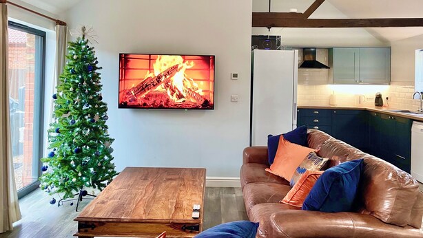 A modern living room with a fireplace, a Christmas tree, and a kitchen in the background. - The Long Barn, Brick Kiln Barns, Dilham - Sleeps 6 in Luxury - Dog Friendly (Dilham)