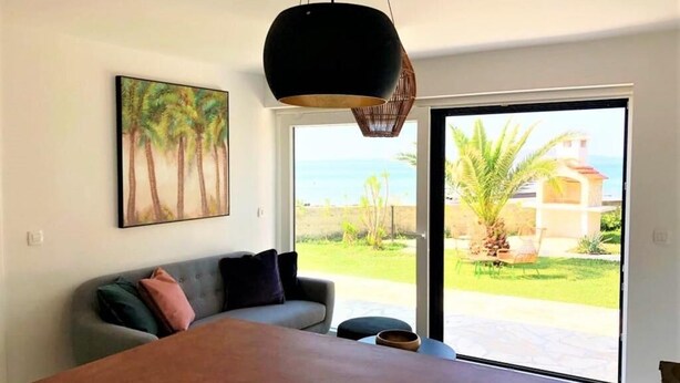 A modern bedroom with a large bed, a sofa, and a view of a garden and ocean through a sliding glass door.