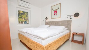 A modern bedroom with a wooden bed, a window with a view of trees, a wall-mounted artwork, and a small bedside table.