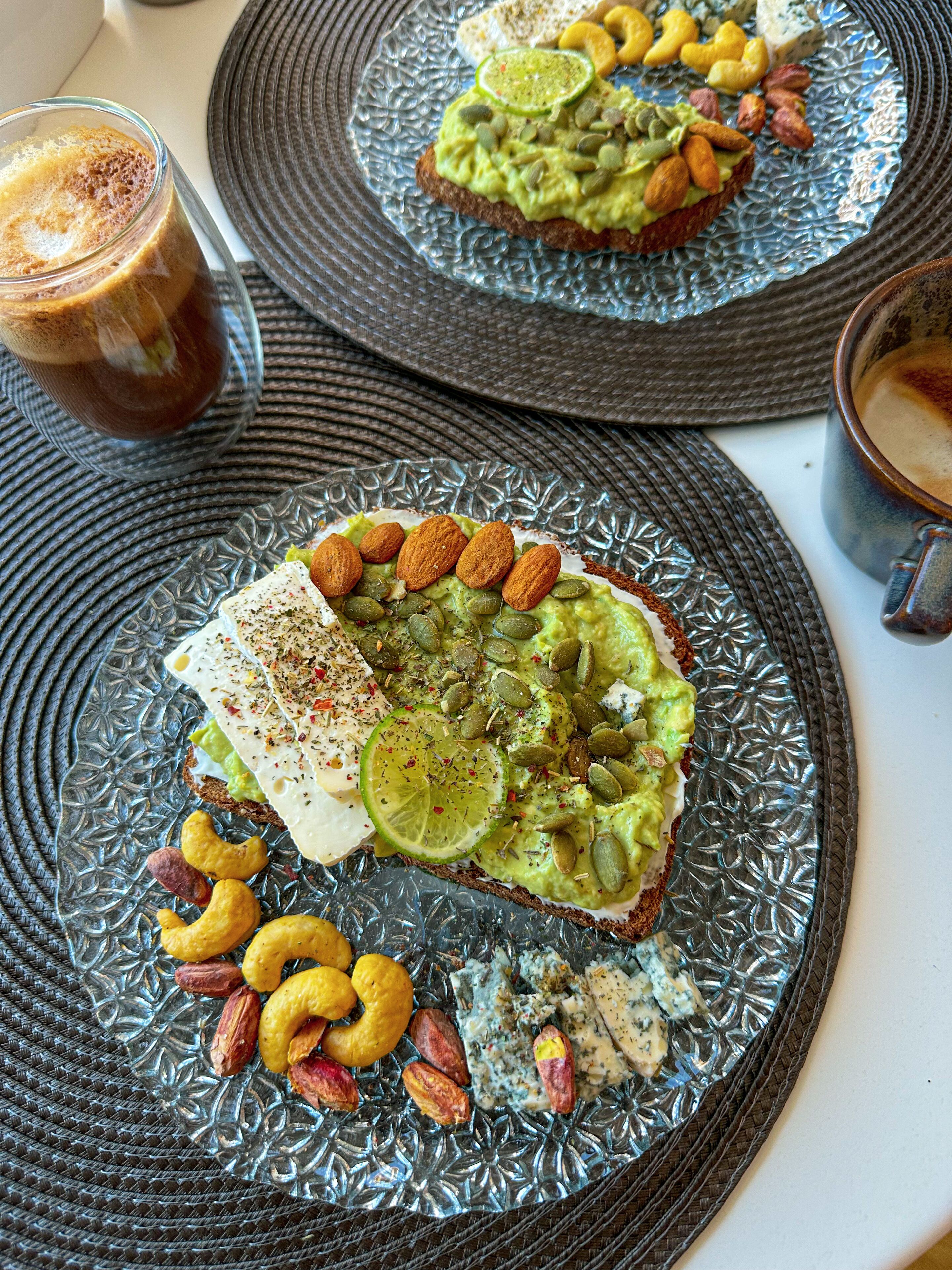 A breakfast setting with two plates of avocado toast, nuts, and a glass of coffee.