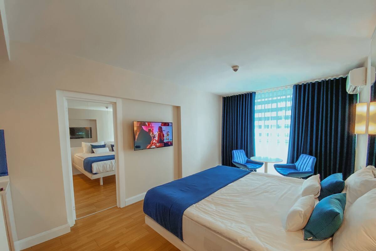 A hotel room with a large bed, a television, a desk with two chairs, and a view of the city through the window.