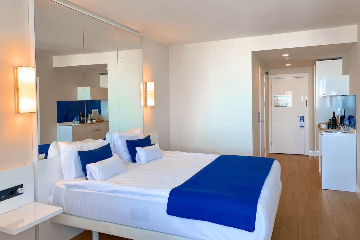 A modern hotel room with a large bed, bedside tables, a mirror, a wardrobe, and a kitchenette.
