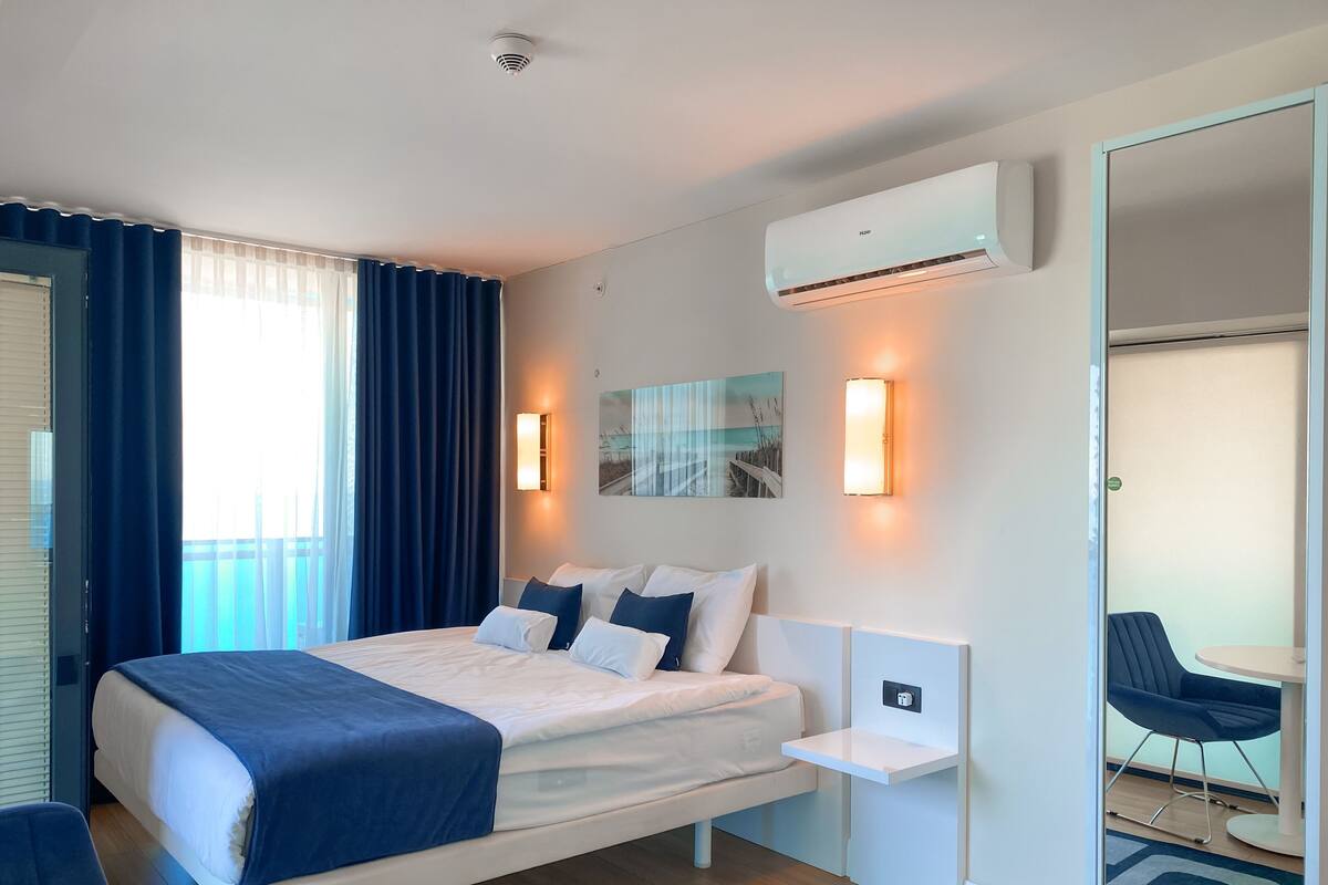 A modern hotel room with a bed, bedside tables, an air conditioning unit, a wall-mounted light, and a view of the outside.