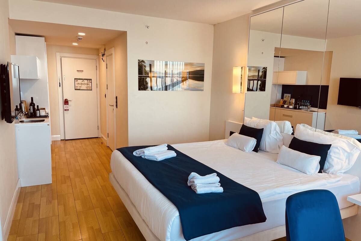 A hotel room with a large bed, a desk, a chair, a mirror, and a kitchenette.