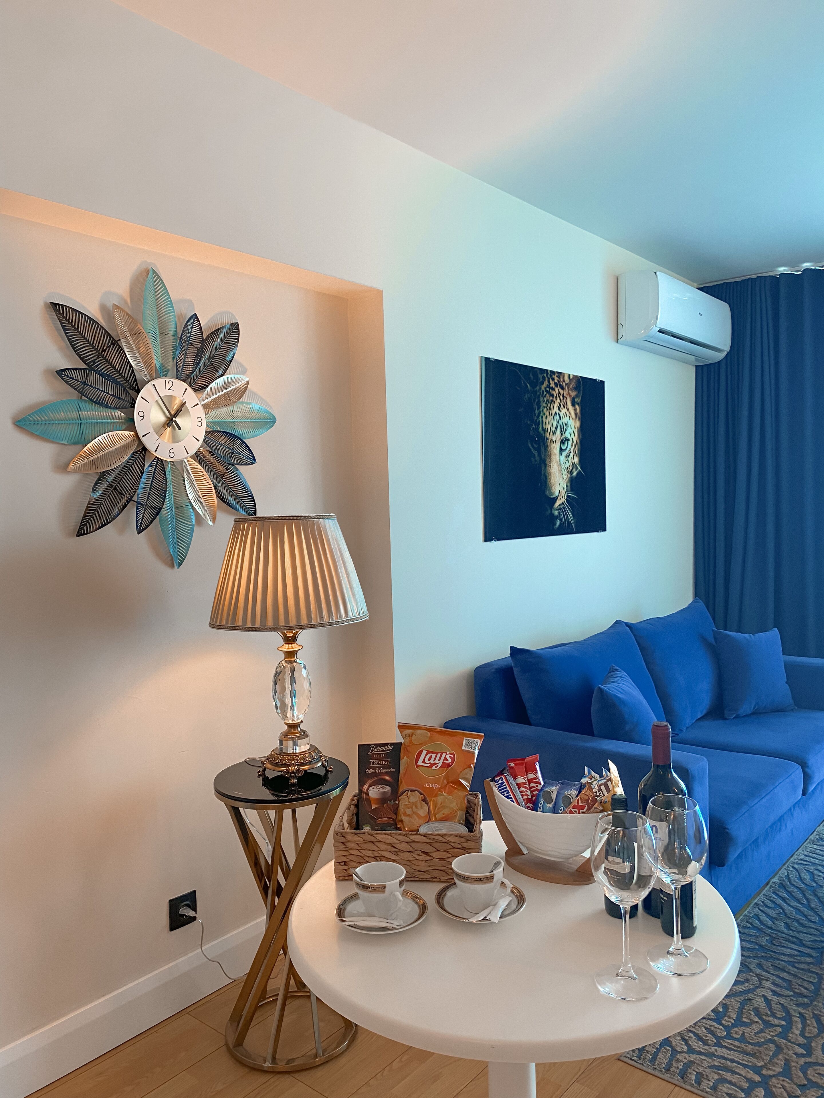 A modern living room with a round white table, a decorative metal clock, a lamp, a blue sofa, a framed artwork, and a basket of snacks.