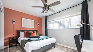 Ein Schlafzimmer mit Bett, Schreibtisch, Deckenventilator, Fenster mit Vorhängen und Blick auf ein Gebäude.