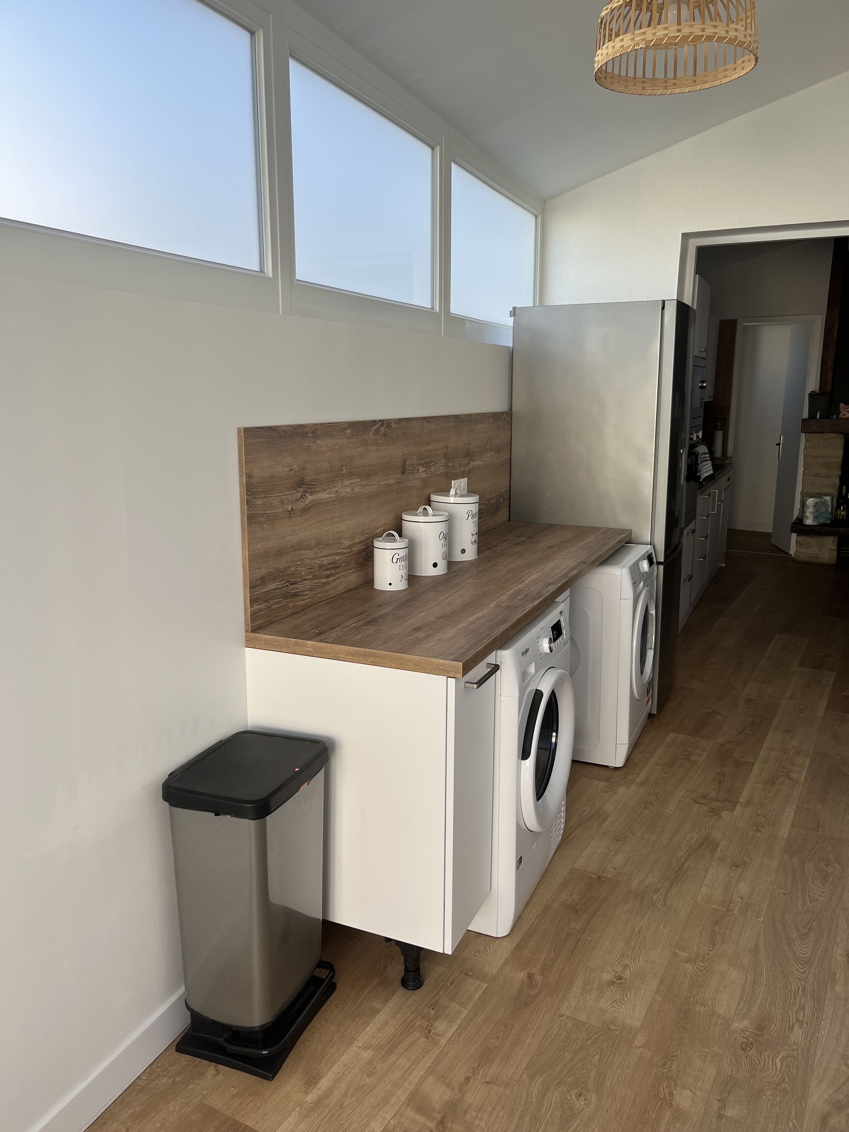A laundry area with a washing machine, dryer, and trash can. There are three windows providing natural light.