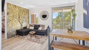 A modern living room with a sectional sofa, a coffee table, and a wooden dining table. There is a large window with a view of greenery outside. - 12 Calming Coral Beach Beautiful 2BRM (Scarborough)