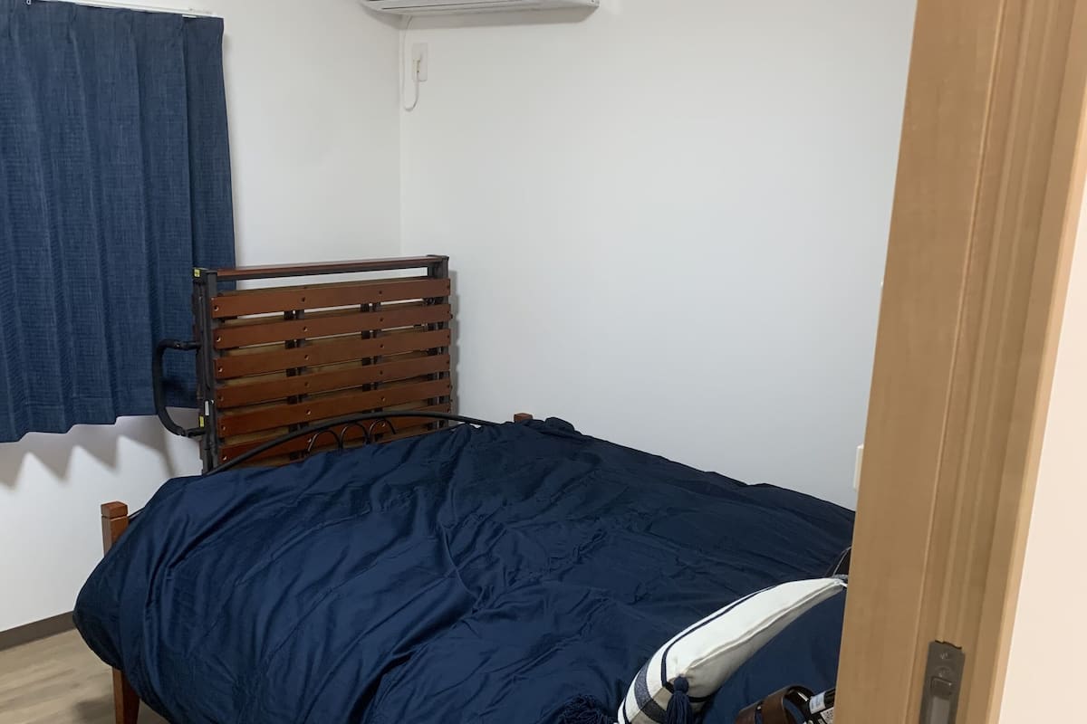 A bedroom with a wooden bed frame, a blue bedspread, a wooden nightstand with a glass bowl, and a white air conditioning unit on the wall.