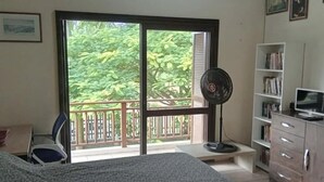 A room with a bed, a desk, a chair, a fan, a bookshelf, and a balcony with a view of greenery. - Comfortable house in Florianópolis |Moçambique Beach (Florianópolis)