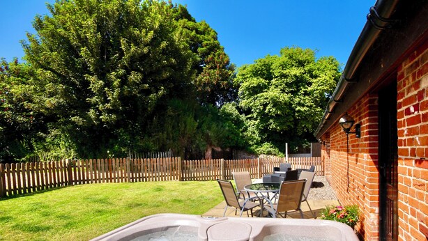 A hot tub with a wooden deck and chairs, surrounded by a brick wall and a wooden fence. - Dog and family friendly barn conversion with fully enclosed garden and hot tub (Dilham)