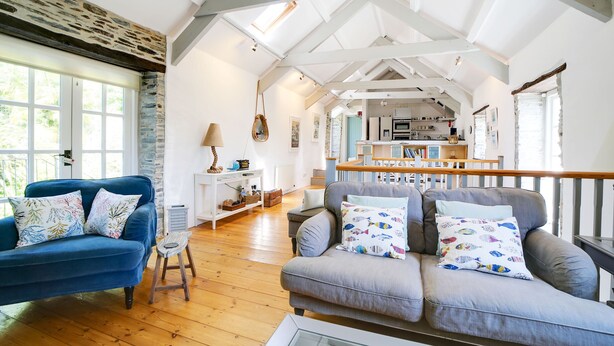 A spacious living area with a high ceiling, wooden beams, a blue sofa, a grey sofa, and a kitchen in the background. - Stone Barn is a stunning barn in an idyllic hamlet, sleeps 6, games room, + dogs (Lanteglos)