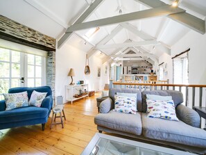 A spacious living area with a high ceiling, wooden beams, a blue sofa, a grey sofa, and a kitchen in the background.