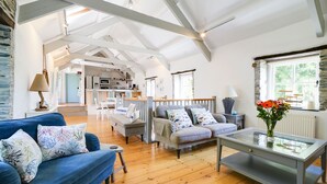 A spacious living area with a blue sofa, a grey sofa, and a glass coffee table. A kitchen is visible in the background, and there is a window with a view of greenery. - Stone Barn is a stunning barn in an idyllic hamlet, sleeps 6, games room, + dogs (Lanteglos)
