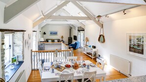 A spacious loft-style living area with a high ceiling, exposed wooden beams, and a cozy seating area with a sofa and armchairs. A dining table is set with plates, cups, and a fruit bowl. - Stone Barn is a stunning barn in an idyllic hamlet, sleeps 6, games room, + dogs (Lanteglos)