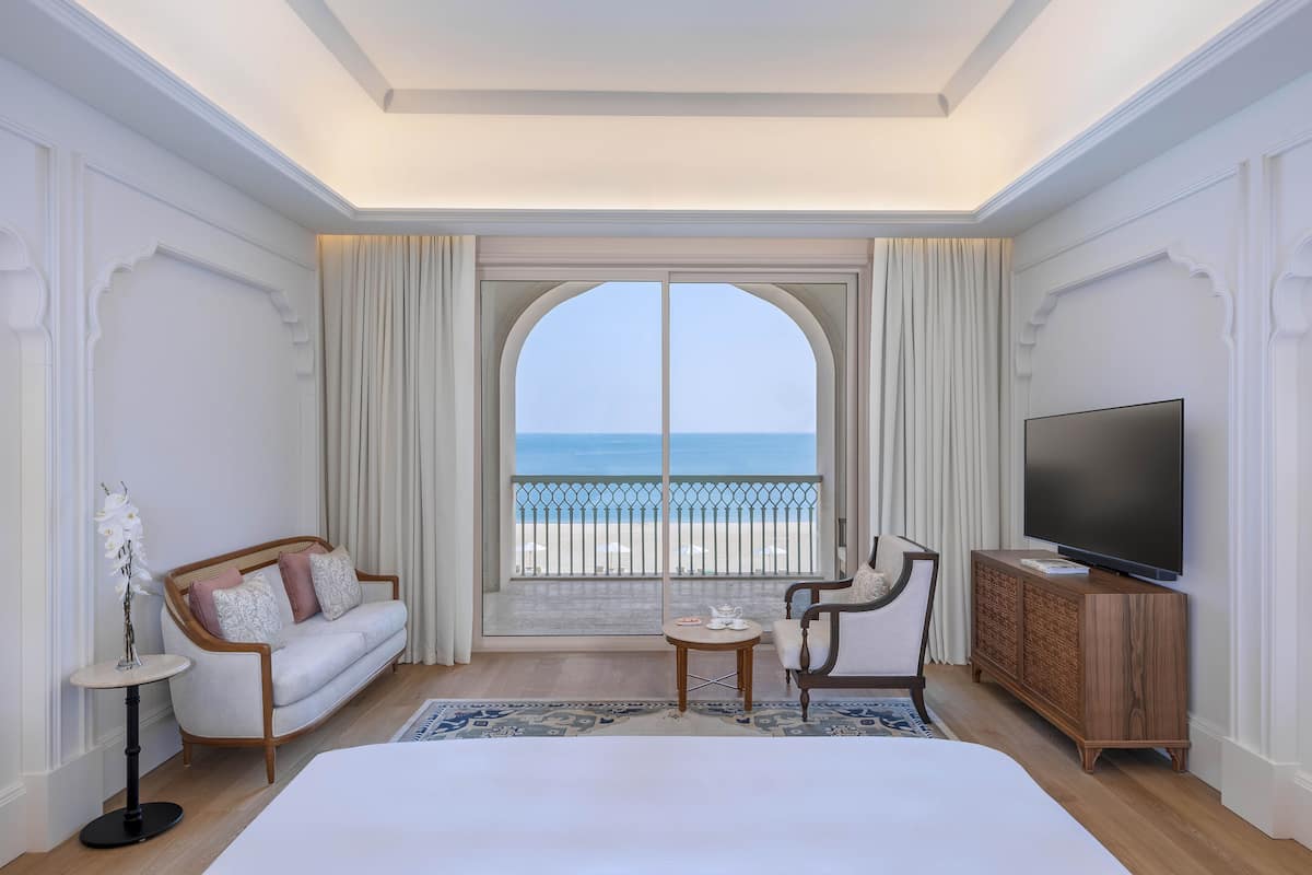 Habitación de hotel con una cama grande, televisión, sofá, sillón, una mesita y vista a la playa.