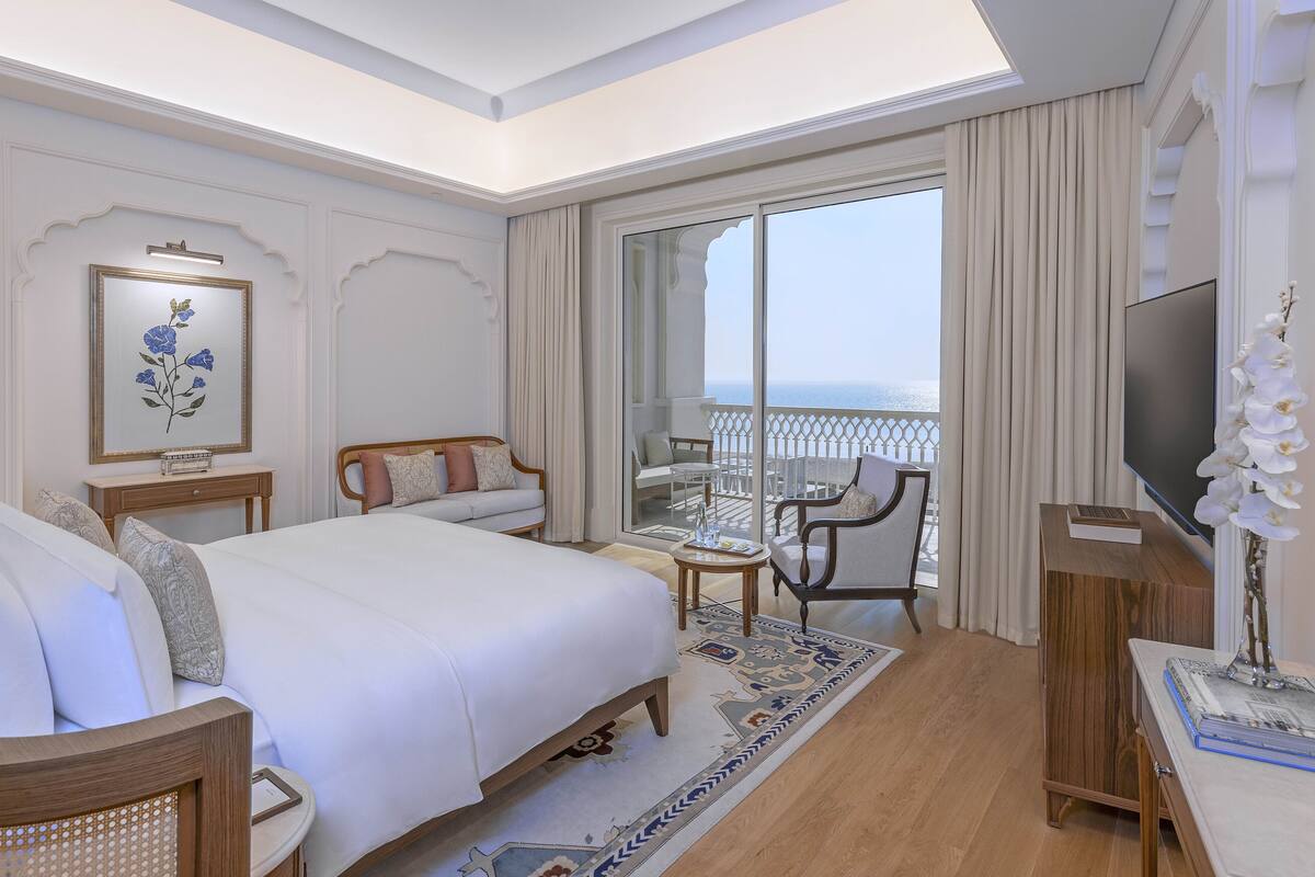 Un cuarto de hotel con una cama grande, una zona de estar con una mesa y sillas, un televisor y un balcón con vista al mar.