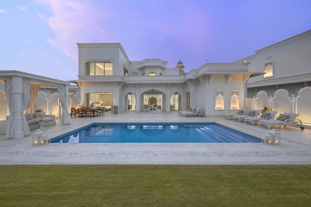 Un edificio grande y blanco de varios pisos con piscina, mobiliario exterior y césped bien cuidado.