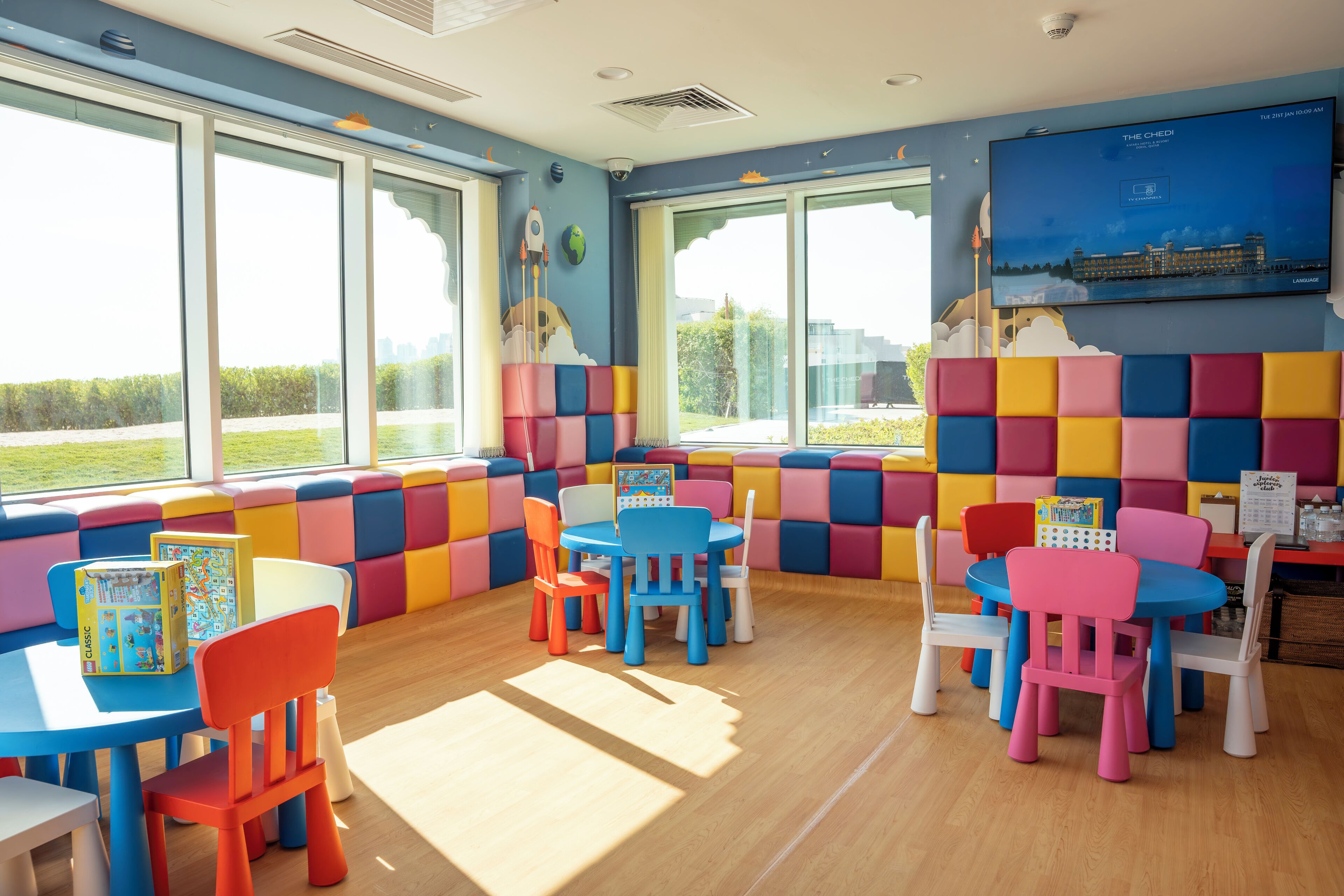 Un espace de jeux pour enfants avec des sièges et des tables rembourrés aux couleurs vives, de grandes fenêtres donnant sur un paysage verdoyant et un écran de télévision affichant le nom de l’hôtel « The Chedi ».