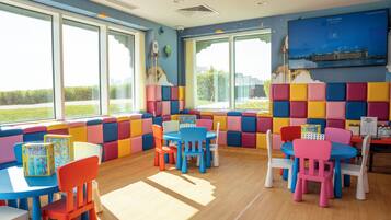 Un espace de jeux pour enfants avec des sièges et des tables rembourrés aux couleurs vives, de grandes fenêtres donnant sur un paysage verdoyant et un écran de télévision affichant le nom de l’hôtel « The Chedi ».