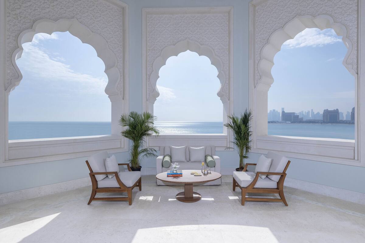 Habitación con sofá blanco, dos sillas de madera, una mesita de centro y dos plantas en macetas. Amplias ventanas con diseños de arco ofrecen vistas al mar y al horizonte de la ciudad.