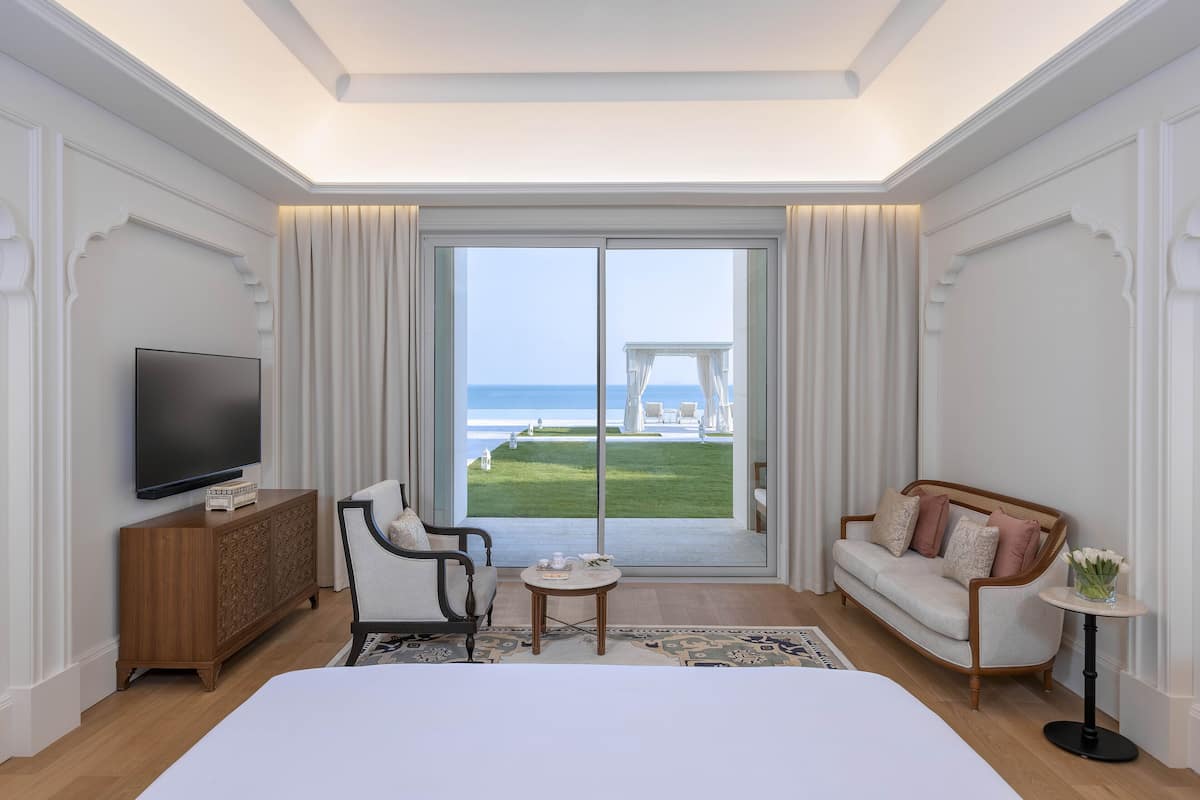 Habitación de hotel con una cama grande, televisión, sofá, una mesita y vistas a la playa a través de puertas correderas amplias.