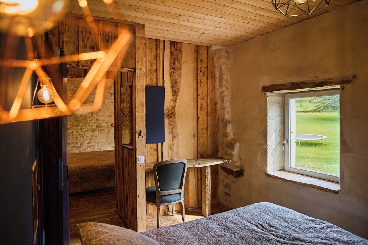 A rustic room with a wooden wall, a small desk, a chair, a bed, and a window with a view of a green field.