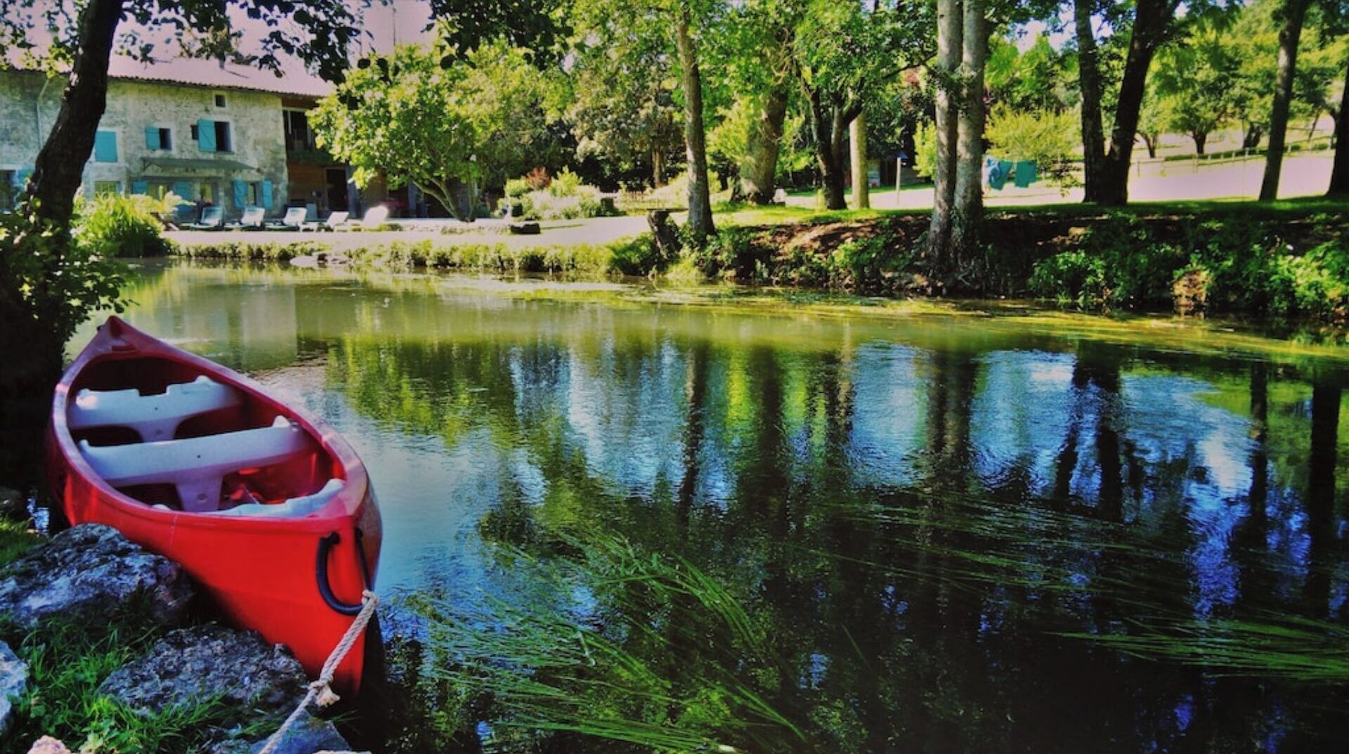 A calm lake with a red boat tied to a dock, surrounded by trees and a building in the background.