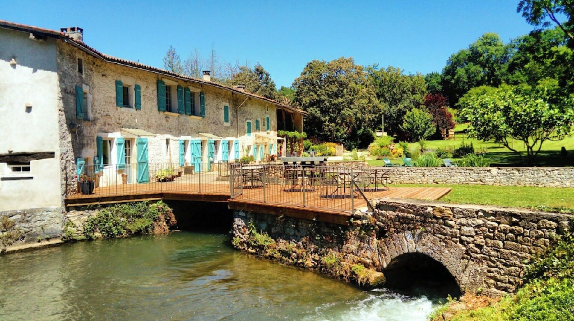 A rustic stone bridge over a river leading to a building with multiple windows and a wooden deck.
