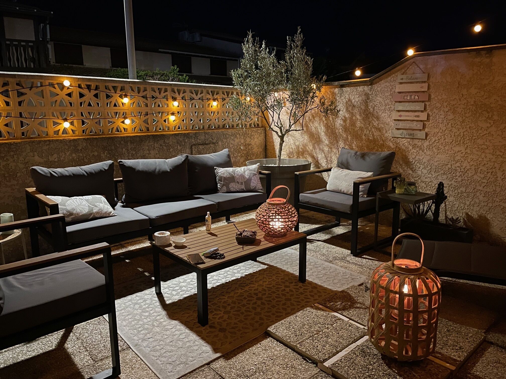 Un espace extérieur aménagé avec un canapé, des fauteuils et une table basse. On y trouve également des guirlandes lumineuses décoratives et une lanterne.