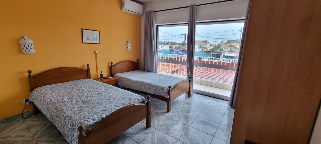A room with two wooden beds, a window with a view of buildings, and a balcony with a railing.