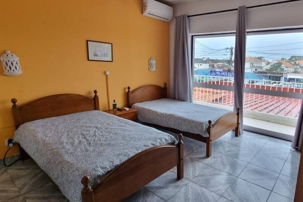 A room with two wooden beds, a window with a view of buildings, and a balcony with a railing.