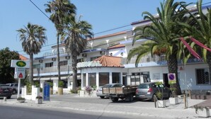 Un edificio de varios pisos con un restaurante llamado "DENGIAL" en la planta baja. Palmeras en la parte delantera y algunos coches estacionados afuera.