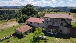 Ein Steinhaus mit rotem Ziegeldach, einem kleinen Anbau und Blick auf sanfte Hügel und Bäume im Hintergrund.