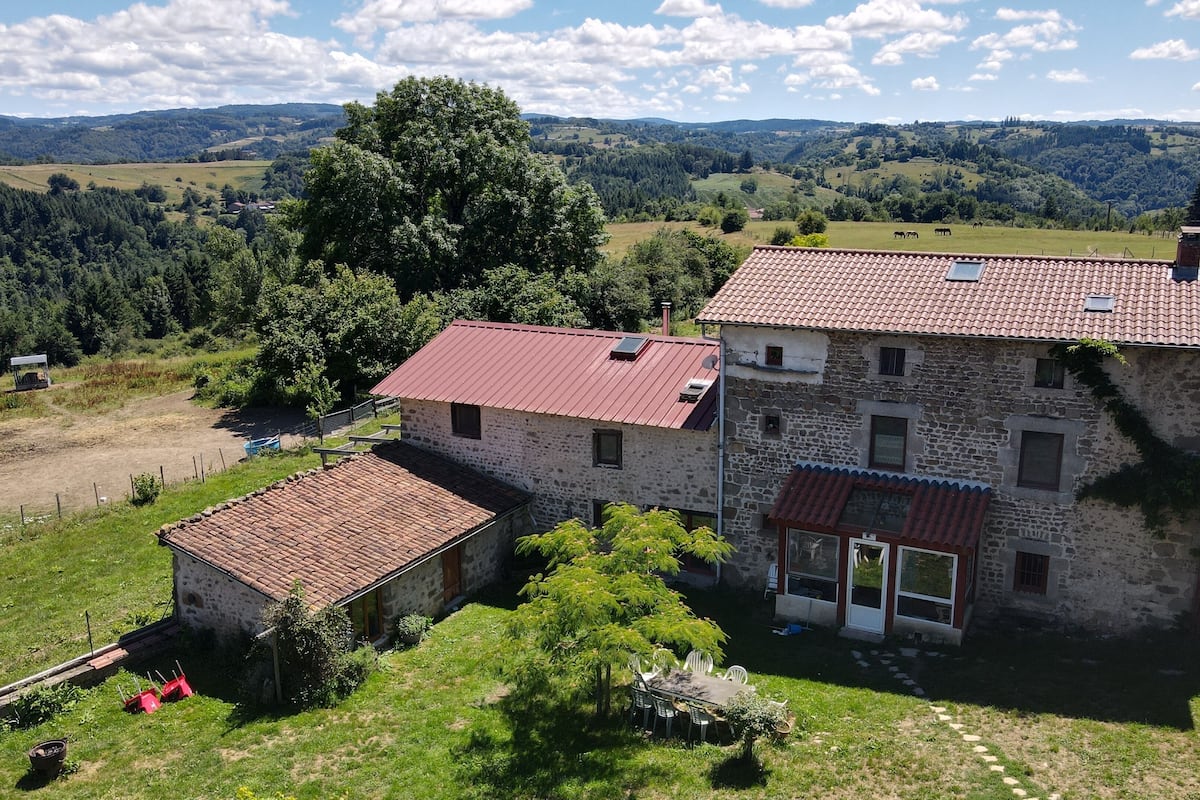 A stone farmhouse with a red-tiled roof, a small attached structure, and a view of rolling hills and trees in the background.
