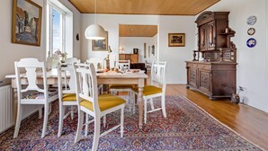 Un comedor con una mesa de madera, sillas blancas con cojines mostaza, un tapete estampado y un aparador de madera con objetos decorativos.